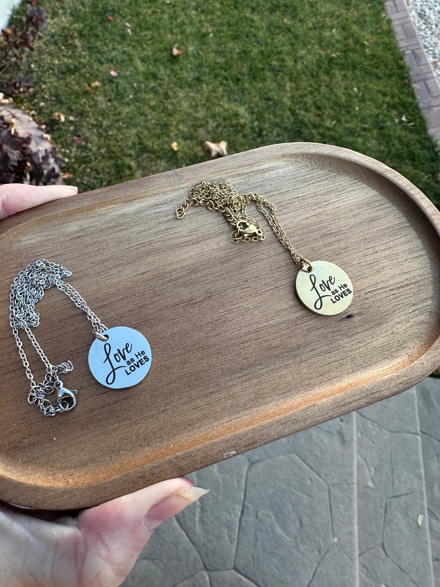 Two necklaces on a wooden tray with 'Love as He loves' tag, held by a hand outdoors.