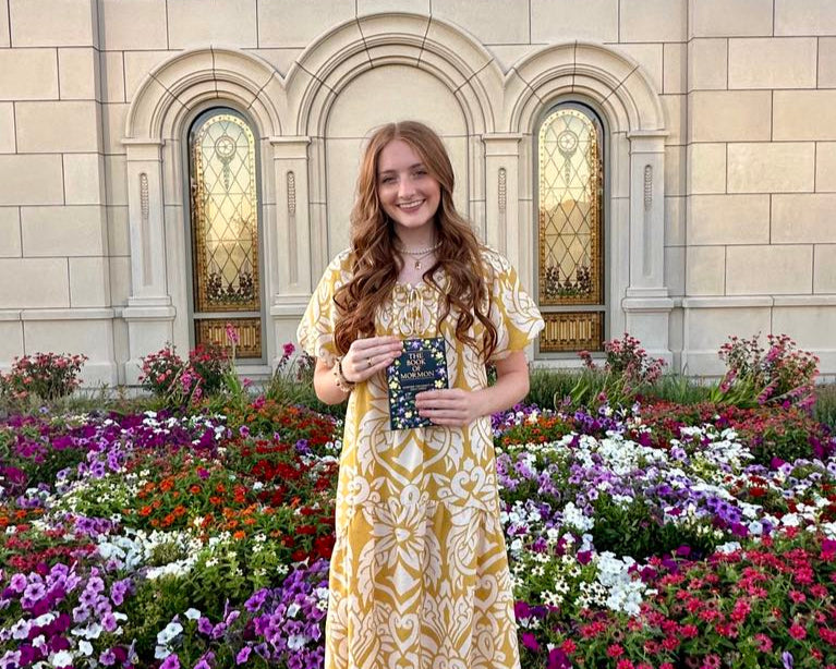 Young woman in a yellow dress holding the Book of Mormon in front of a garden