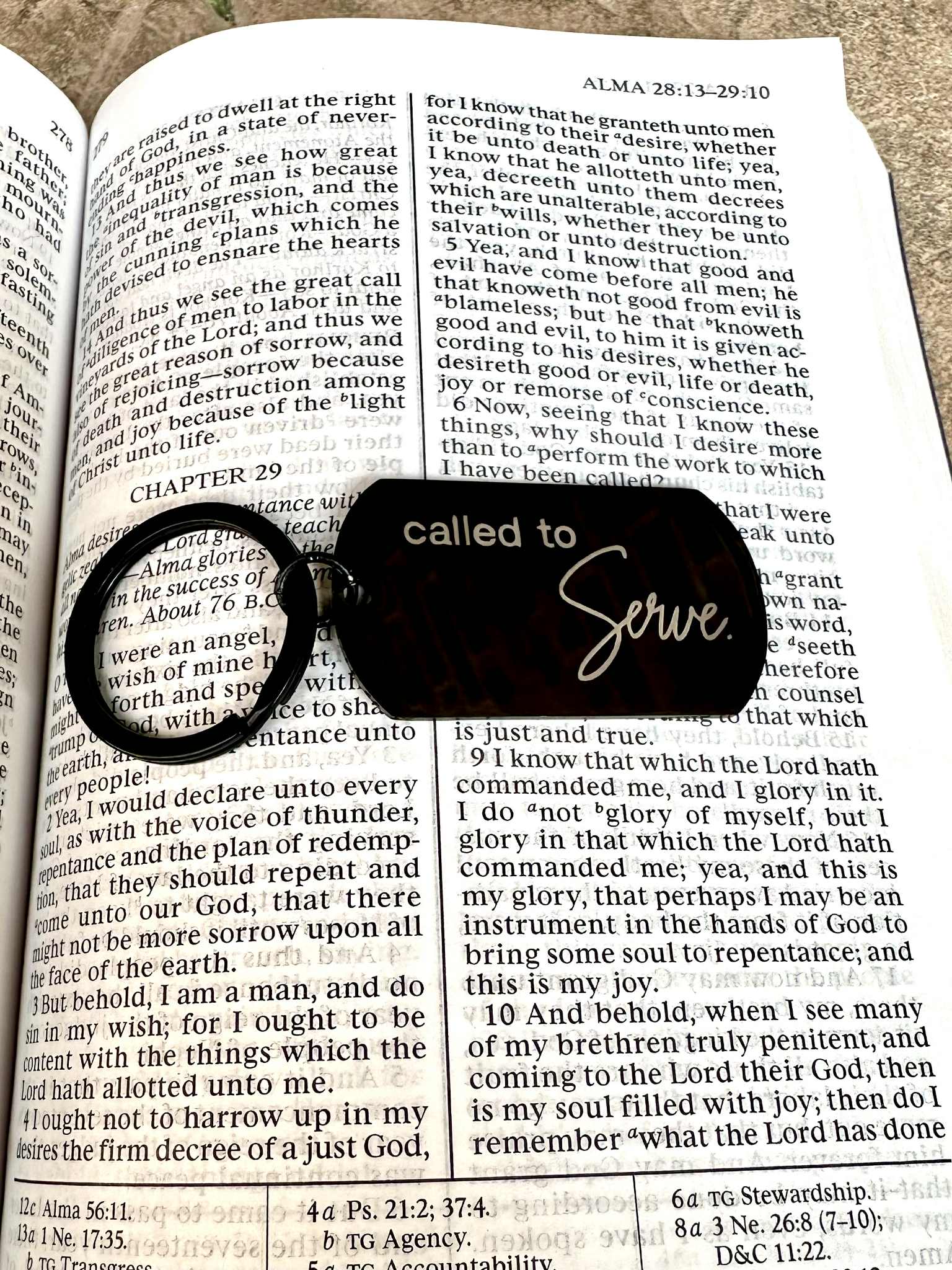 Black stainless steel tag engraved with the words "Called to Serve." in silver. An open page of the Book of Mormon is used as the background.