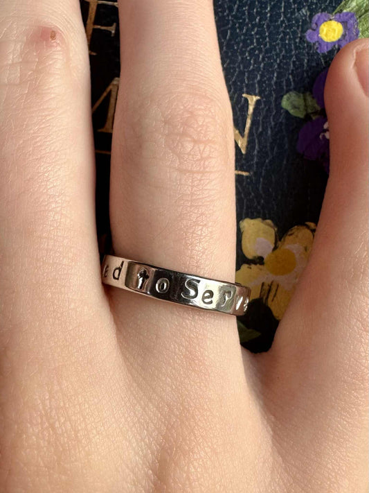 Silver ring with engraved text that says "called to serve" on a finger against a floral background