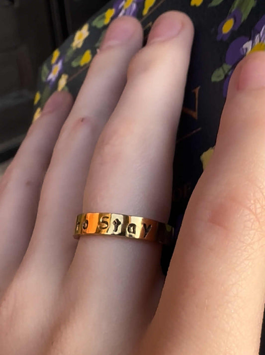 Gold ring with the engraved text "called to stay" on a person's finger against a floral-patterned background