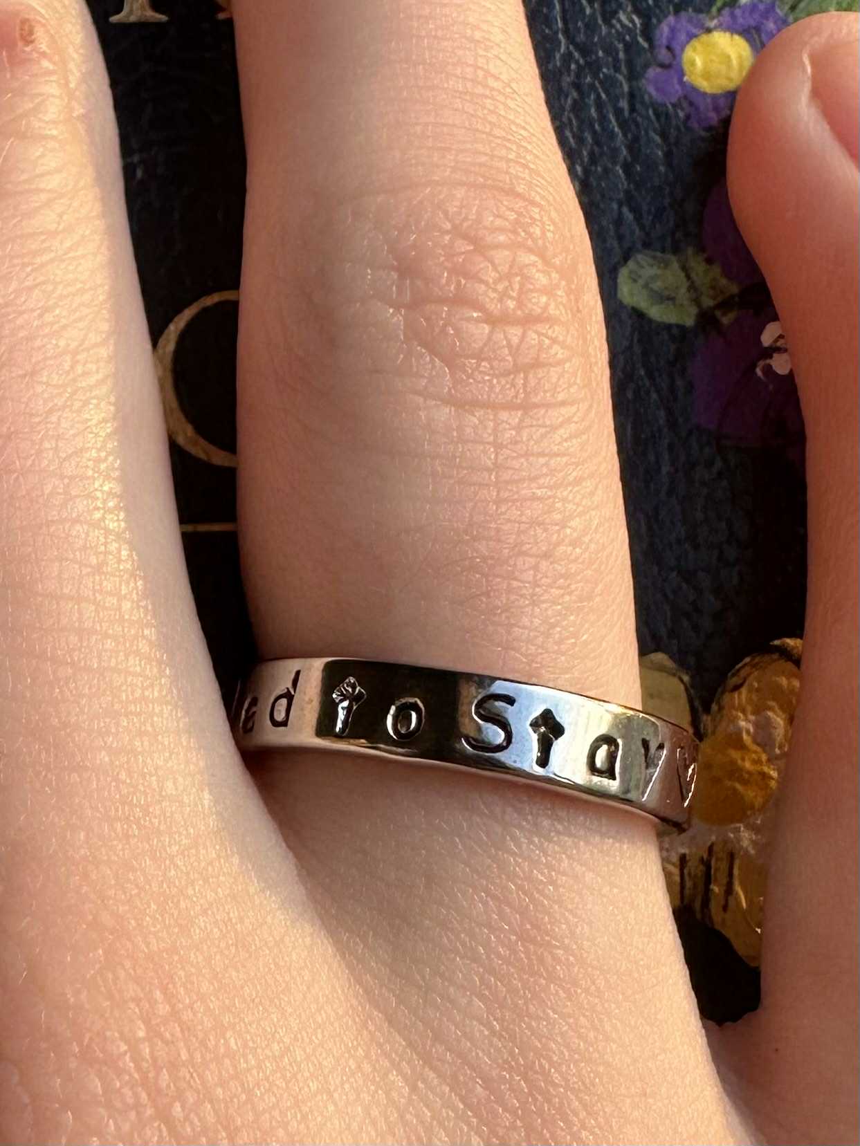 Silver ring with engraved text that says "called to stay" on a person's finger against a floral background
