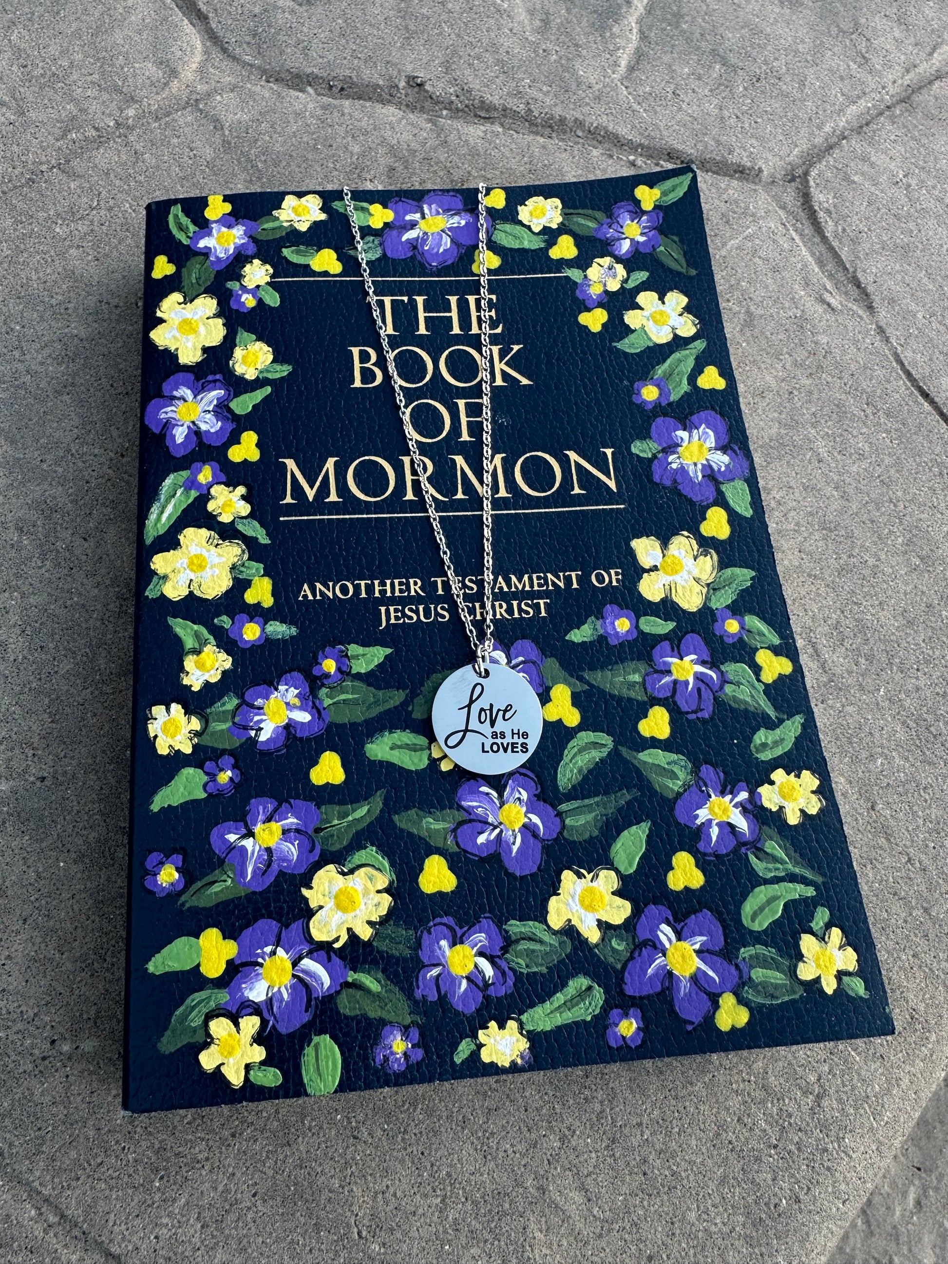 Book of Mormon with floral design on a textured surface with a silver love as He loves necklace on top