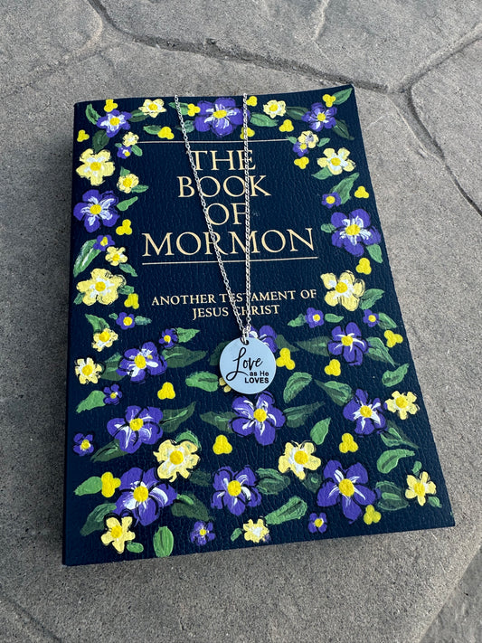 Book of Mormon with floral design on a textured surface with a silver love as He loves necklace on top