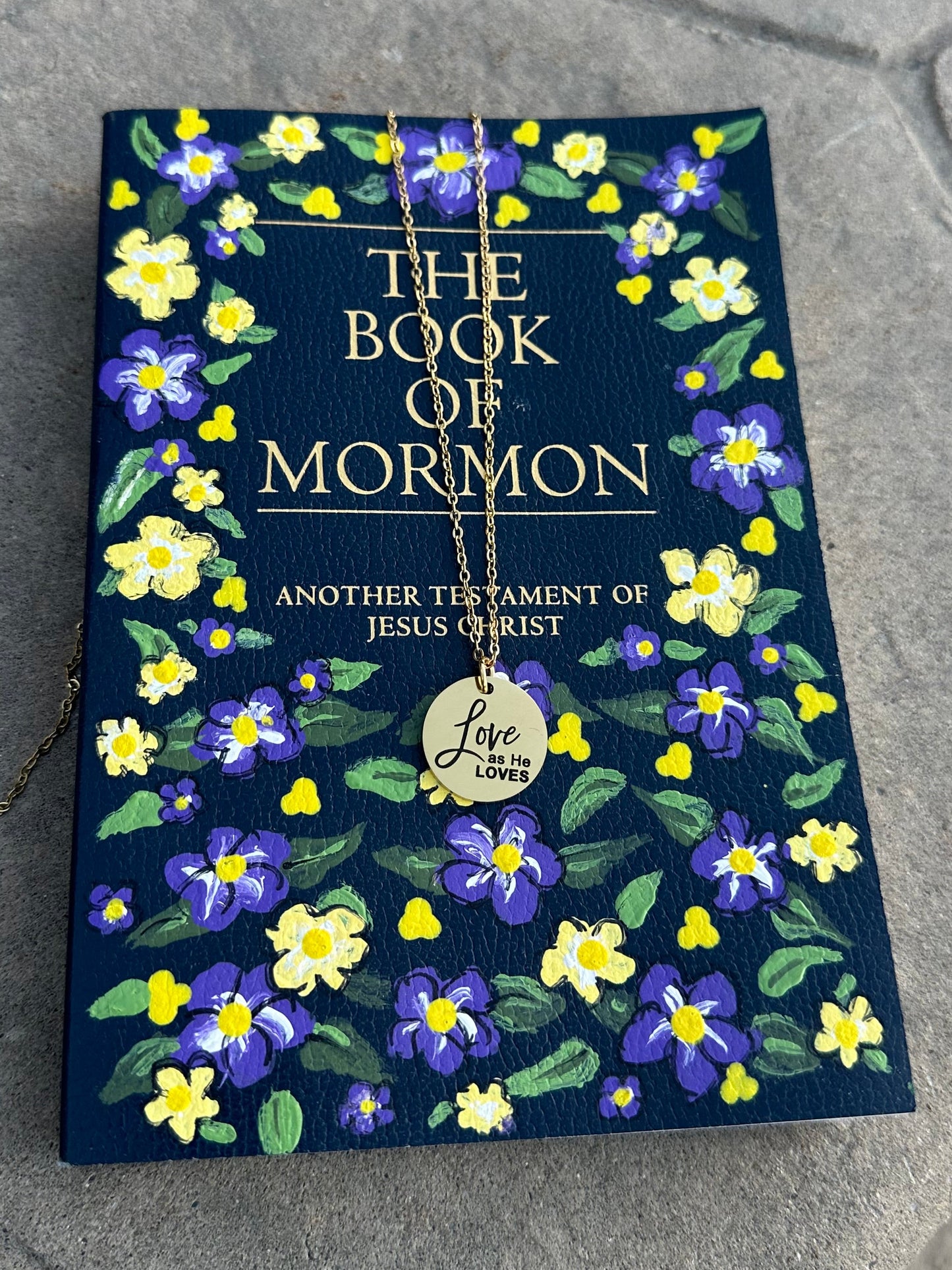 Book of Mormon with floral design on a textured surface with a gold love as He loves necklace on top