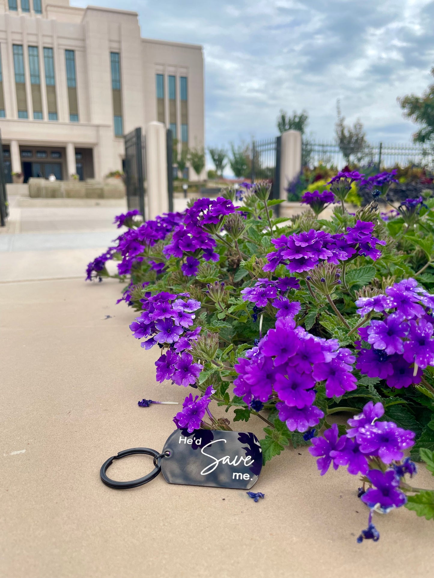Black 'He'd save me' keychain next to purple flowers with the temple in the background
