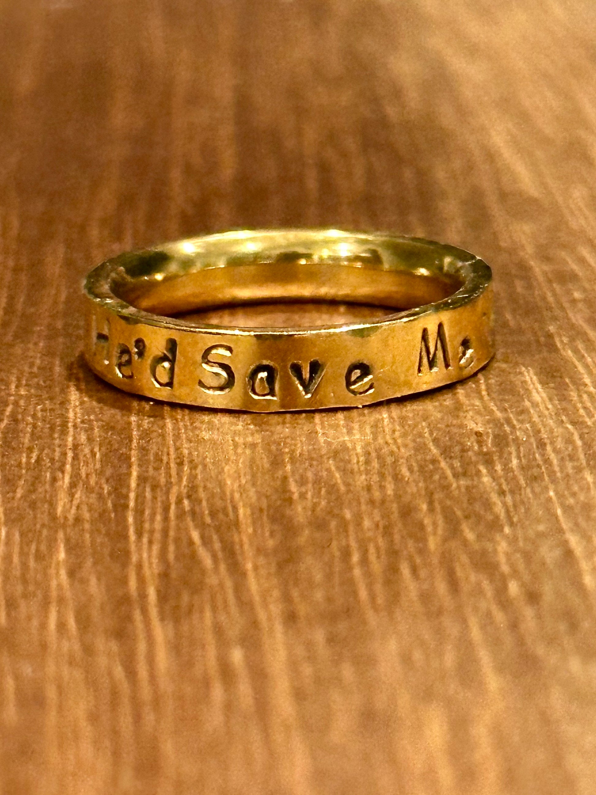 Gold ring that says 'He'd Save Me on a wooden surface
