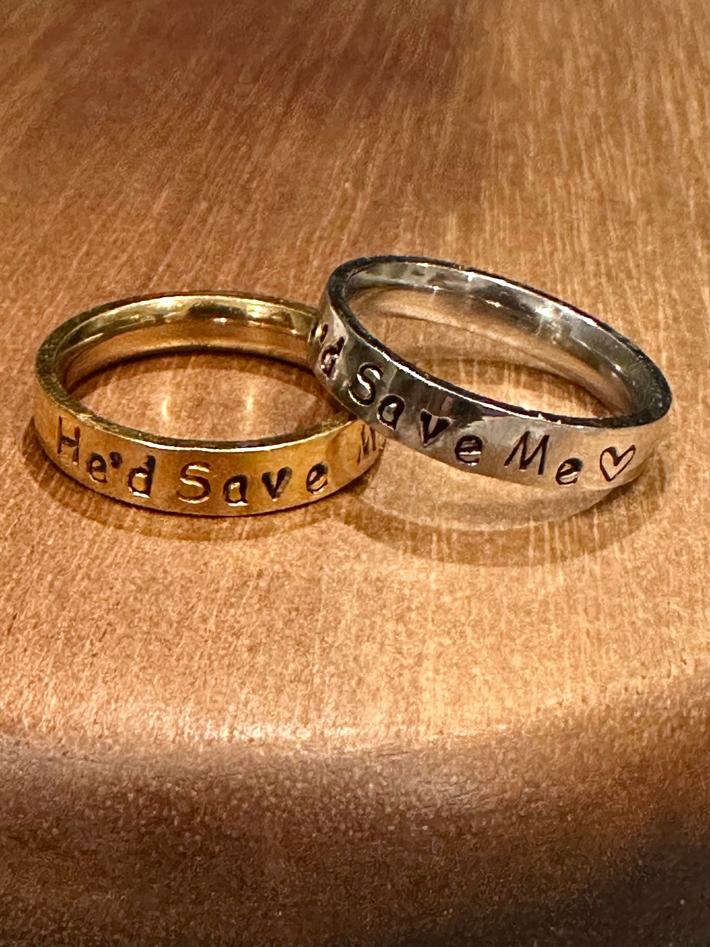 Two rings, one gold and one silver, with 'He'd Save Me' on it on a wooden surface.