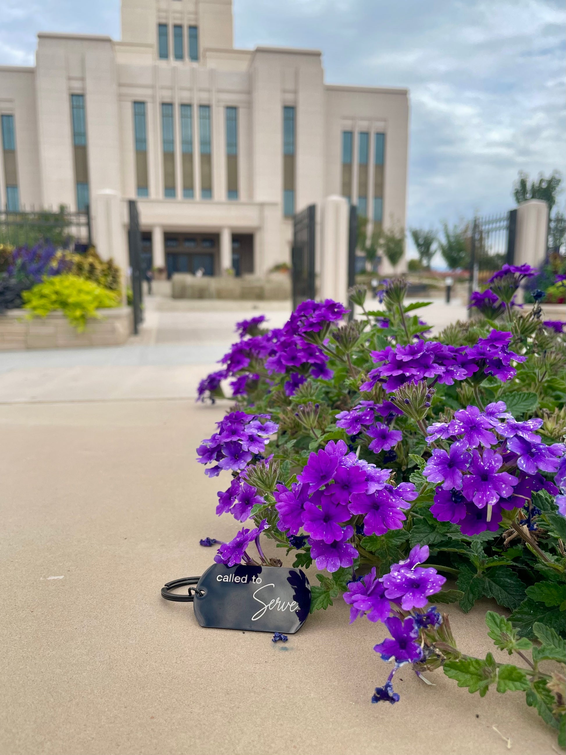 Black 'Called to serve' keychain next to purple flowers with the temple in the background