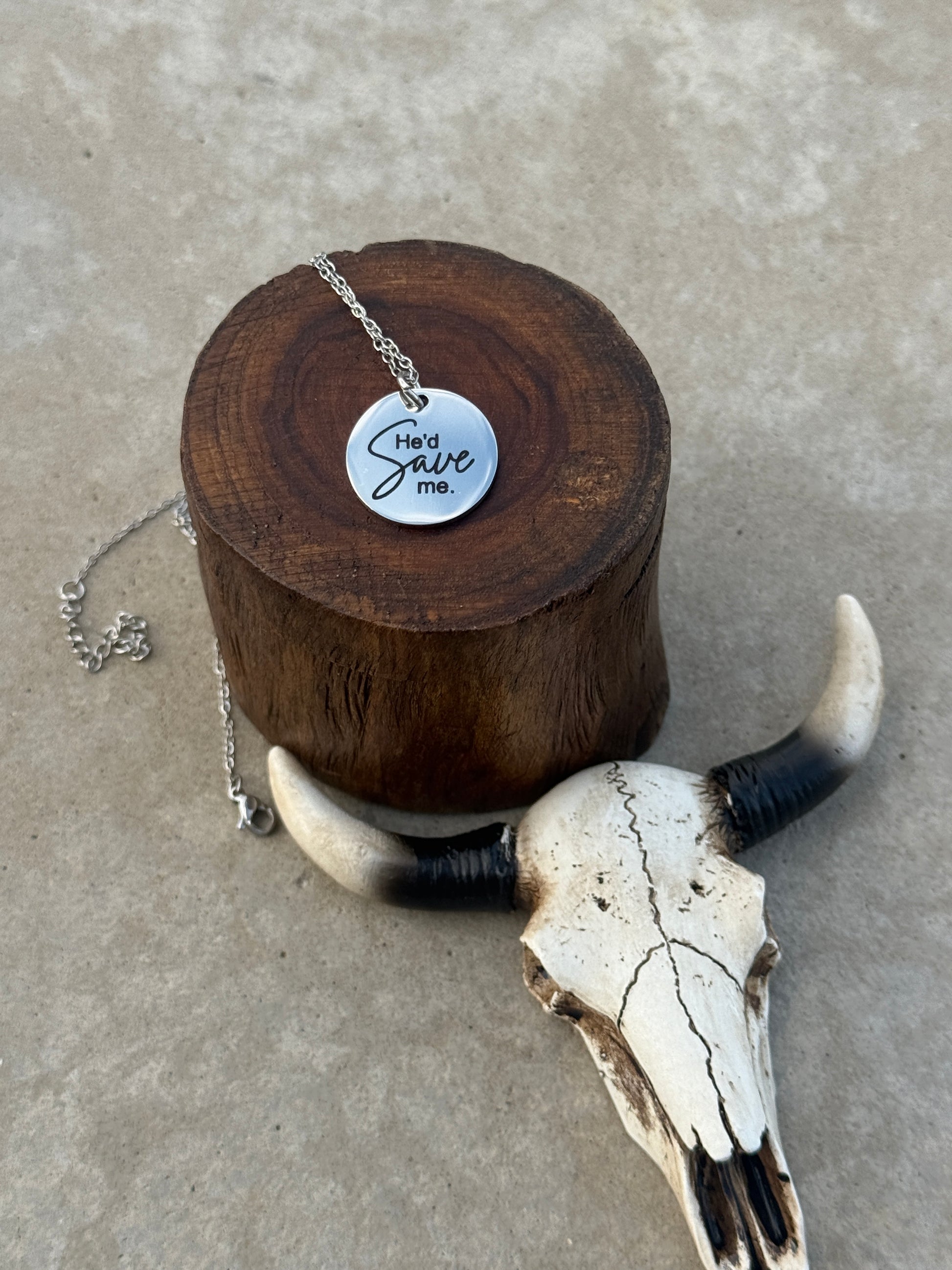 Silver He'd Save Me necklace on a wooden stump with a cow skull next to it. 